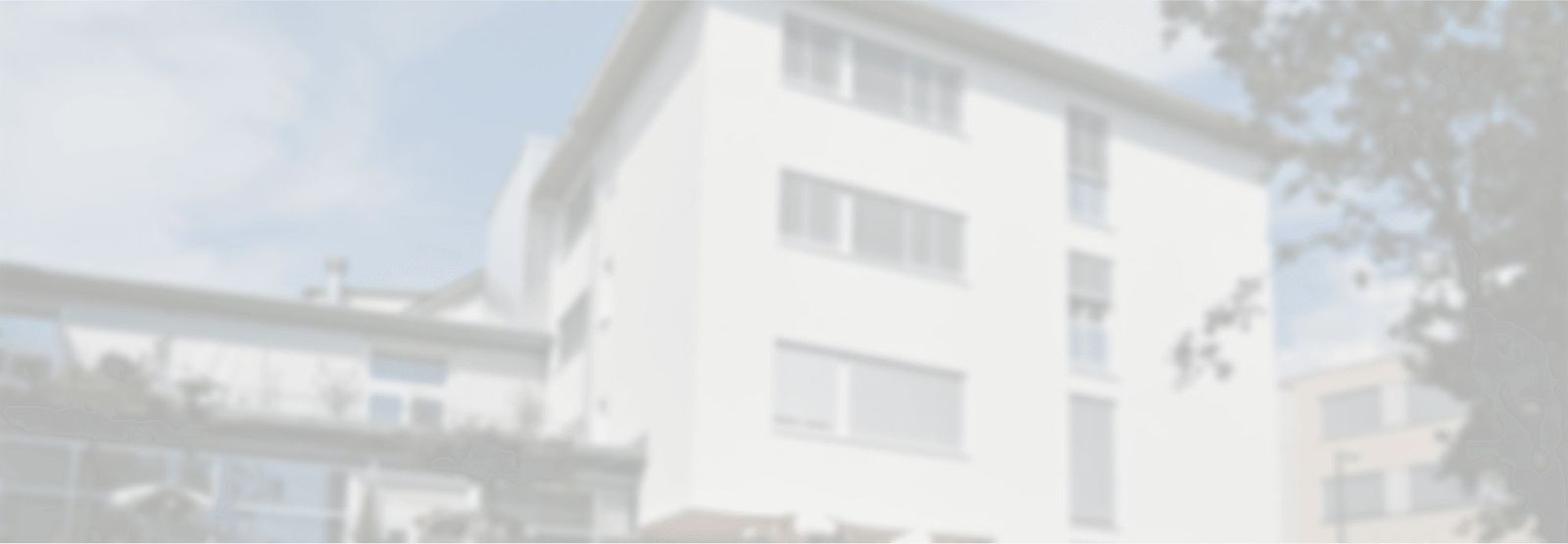 Mehrstöckiges Wohngebäude mit weißen Fassaden und mehreren Fenstern, umgeben von Bäumen und bewölktem Himmel.