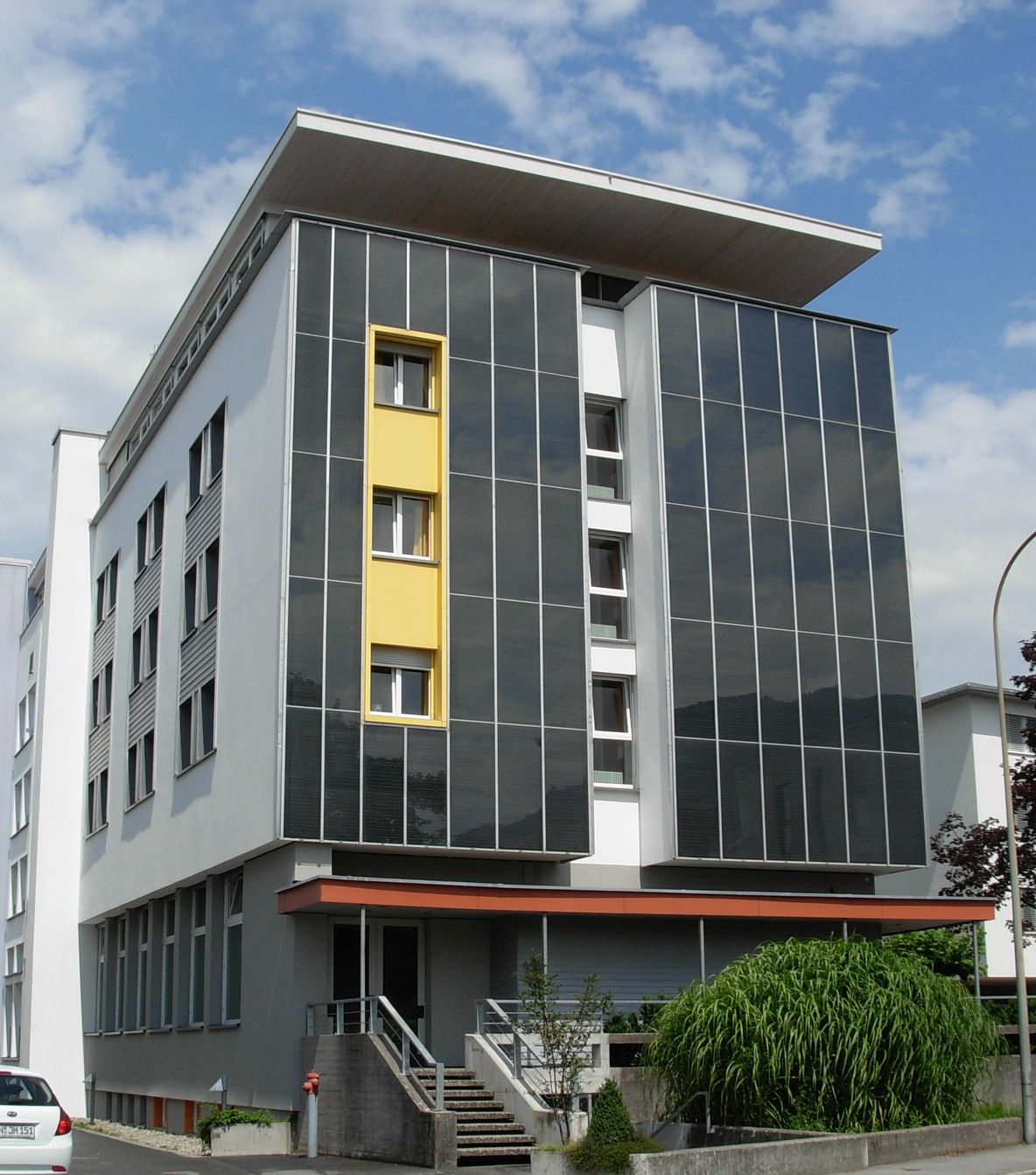 Mehrstöckiges Gebäude mit großen, vertikalen Solarpaneelen an der Fassade und gelb eingerahmten Fenstern.